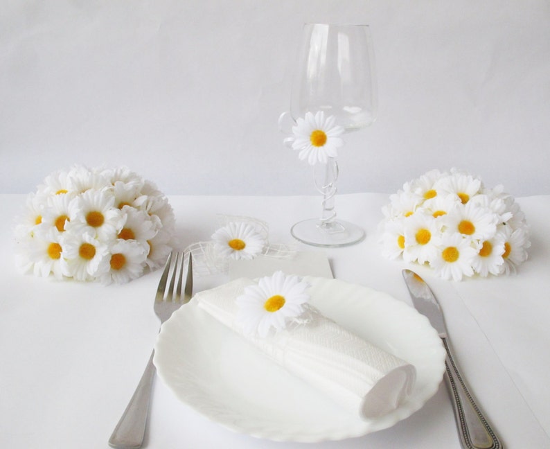 May include: White table setting with a white napkin, a white plate, a fork, a knife, and a glass with a white daisy. There are three white daisy arrangements on the table.