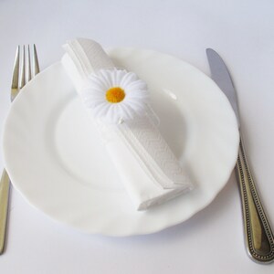 May include: A white napkin ring with a white daisy flower with a yellow centre. The napkin ring is on a white plate with a silver fork and knife.