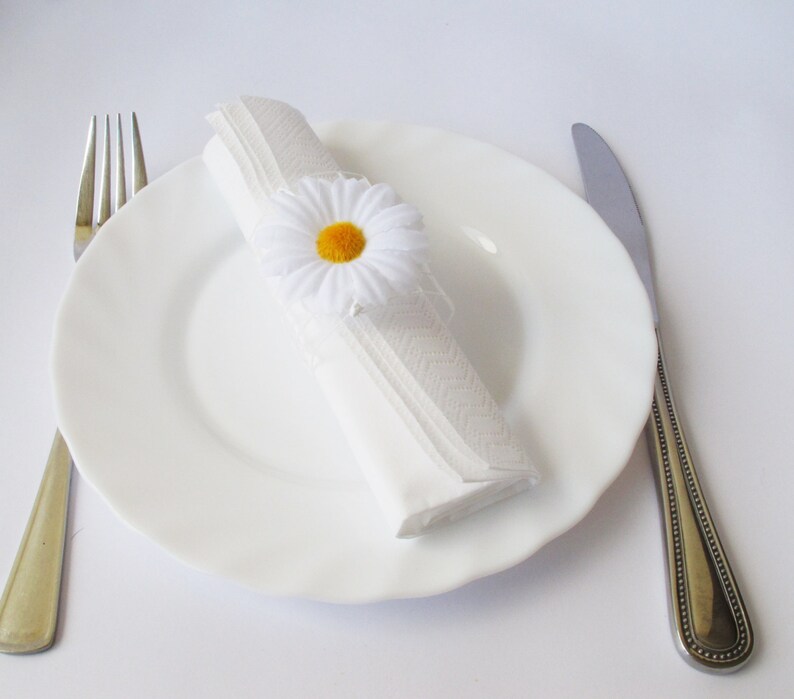 May include: A white napkin ring with a white daisy flower with a yellow centre. The napkin ring is on a white plate with a silver fork and knife.