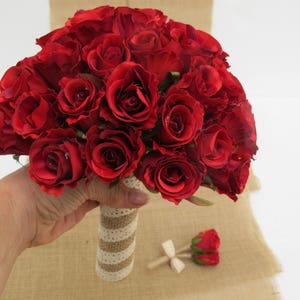 May include: A bouquet of red silk roses with a burlap and lace wrap. The bouquet is held in a hand.