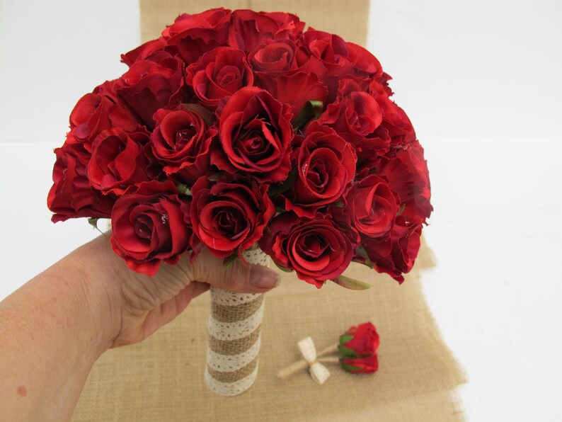 May include: A bouquet of red silk roses with a burlap and lace wrap. The bouquet is held in a hand.