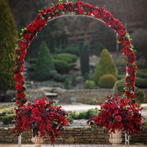 May include: A wedding arch decorated with red roses and greenery, with two matching red rose arrangements in white vases on either side of the arch.