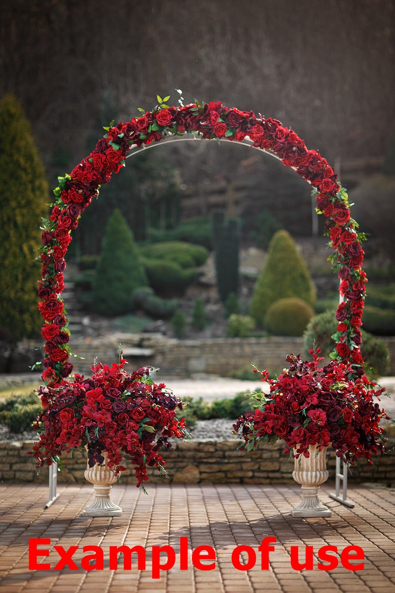 May include: A wedding arch decorated with red roses and greenery, with two matching red rose arrangements in white vases on either side of the arch.