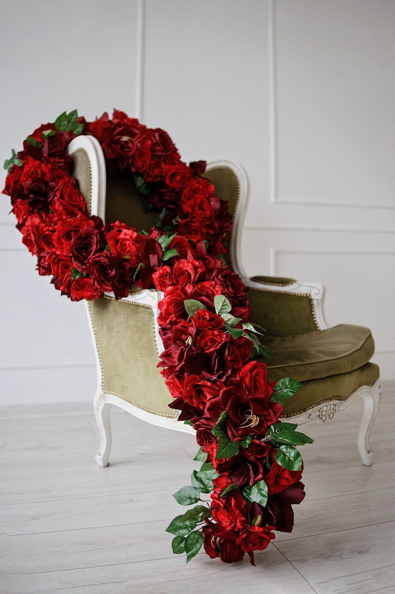 May include: A white armchair with a green velvet seat is decorated with a long garland of red roses. The garland is draped over the back of the chair and cascades down the side.