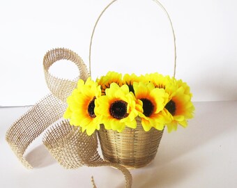 May include: A small, woven basket filled with bright yellow sunflowers with dark brown centres. A decorative burlap ribbon is curled around the basket. The basket has a handle and is set against a white background.