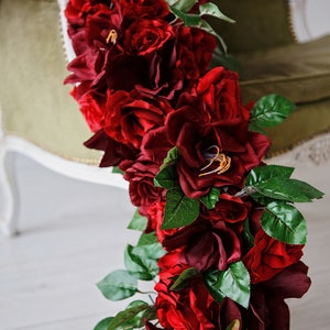 May include: A long garland of deep red artificial roses with green leaves. The garland is draped over a green velvet chair.