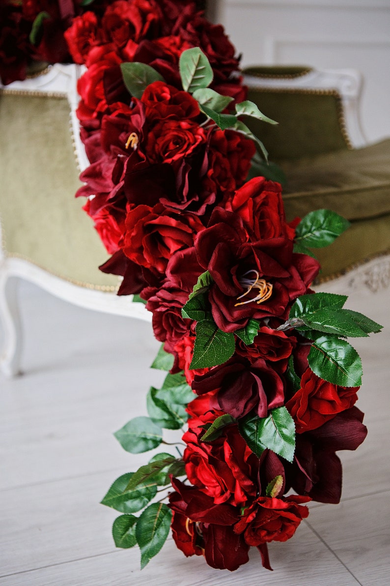 May include: A long garland of deep red artificial roses with green leaves. The garland is draped over a green velvet chair.