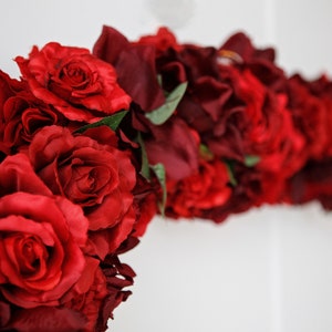 May include: A garland of red and burgundy artificial roses, with green leaves, arranged in a curved shape.