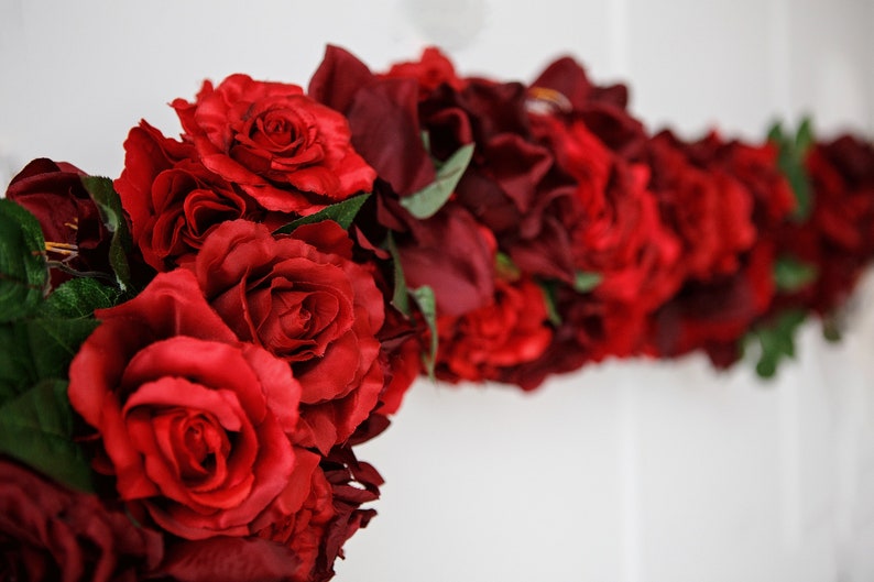 May include: A garland of red and burgundy artificial roses, with green leaves, arranged in a curved shape.