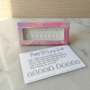 Puede incluir: Una caja de uñas postizas transparentes de forma ovalada con un diseño holográfico rosa y morado. Una guía de tallas con instrucciones y contornos de uñas está al lado de la caja. El texto de la guía dice "Perfect Fit Sizing Guide".