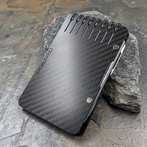 Peut inclure: Un carnet noir en fibre de carbone avec une reliure spirale argentée et le mot "MGEARID" imprimé sur la couverture. Le carnet est doté d'un stylo argenté.