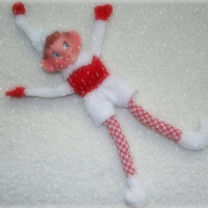 May include: A handmade elf doll with a white body, red accents, and checkered legs. The elf has a red and white outfit, including a red torso, white shorts, and red and white checkered legs. The doll is lying on a white surface.