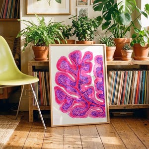 May include: A framed abstract art print featuring a vibrant pink and purple botanical design. The artwork is displayed on a wooden floor, surrounded by plants and a vintage record collection, creating a cozy, artistic atmosphere.