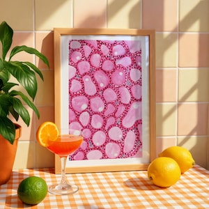 Puede incluir: Una impresión enmarcada con un patrón rosa similar a células. Un cóctel con una rodaja de naranja como adorno está frente a la impresión. Una lima y dos limones están sobre un mantel a cuadros. Una planta en maceta está a la izquierda.