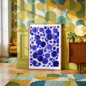 Puede incluir: Una obra de arte enmarcada con un diseño de acuarela de círculos azules y morados de varios tamaños. La obra de arte se exhibe en una habitación con una estética retro.