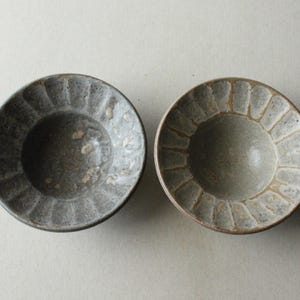 May include: Two handcrafted ceramic bowls with a textured, ridged rim design. One bowl is dark grey, the other is light grey with a tan rim. The bowls are round and shallow, suitable for serving small portions. Each bowl is approximately 15 cm in diameter.