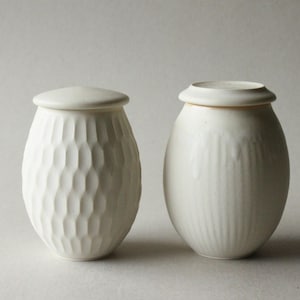 May include: Two off-white ceramic jars with lids. One jar has a textured, wave-like pattern, while the other has vertical lines. Both jars have a rounded, oval shape and are suitable for storing dry goods.