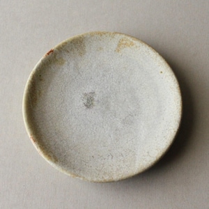 Puede incluir: Un pequeño plato redondo de cerámica con un esmalte texturizado en color blanco roto. La superficie presenta sutiles patrones de grietas y motas de color beige y marrón, con un borde ligeramente irregular y un aspecto terroso.