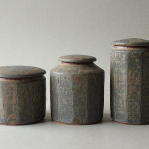 Op de afbeelding: Drie keramische potten met deksels. De potten hebben verschillende maten en een zeshoekig ontwerp. De kleur is een gevlekt grijsgroen met roodbruine accenten. De deksels zijn plat en rond. De potten zijn waarschijnlijk voor opslag.