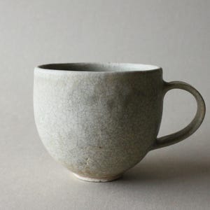 Puede incluir: Una taza de cerámica con cuerpo redondeado y asa curva. El exterior presenta un esmalte texturizado de color gris claro con un patrón de grietas visible en toda la superficie. La base de la taza es ligeramente cónica y muestra un tono beige.