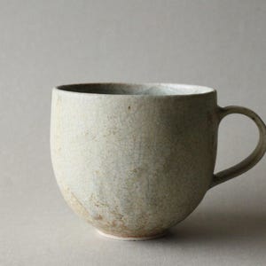 May include: A ceramic mug with a rounded body and a curved handle, featuring a light beige, off-white glaze with a visible crackle pattern throughout the surface and subtle brown earthy tones near the base of the vessel.