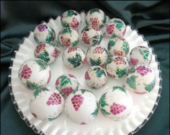 May include: A collection of white, round ornaments with hand-painted grape and leaf designs in shades of purple, green, and gold. The ornaments are arranged on a white, ruffled-edge plate, creating a festive display.