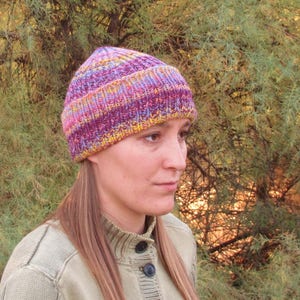 Op de afbeelding: Een vrouw draagt een kleurrijke gebreide muts met paarse, roze, gele en blauwe strepen. De muts is gemaakt van zacht garen en heeft een slouchy pasvorm.