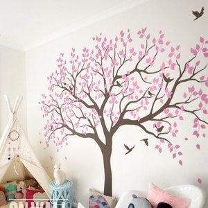 Op de afbeelding: Een roze en bruine boomwanddecoratie met vogels en bladeren. De sticker is op een witte muur in een kamer met een tipi, een knuffeldier en een stoel.