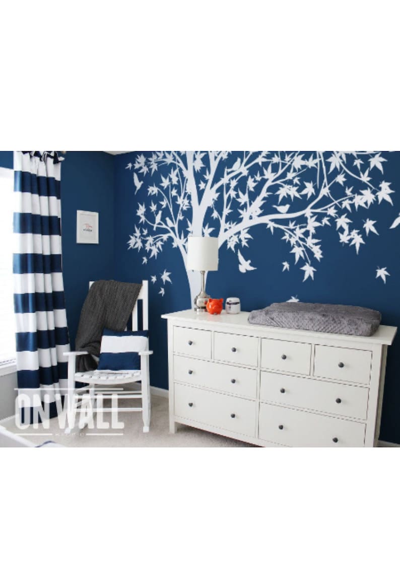 May include: A white tree decal with birds on a dark blue wall. A white dresser with six drawers is in the foreground. A rocking chair with a gray blanket is in the background.