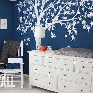 May include: White tree wall decal with birds on a dark blue wall, a white dresser with six drawers, a gray changing pad on top of the dresser, and a rocking chair with a gray throw blanket and a white and blue striped pillow.