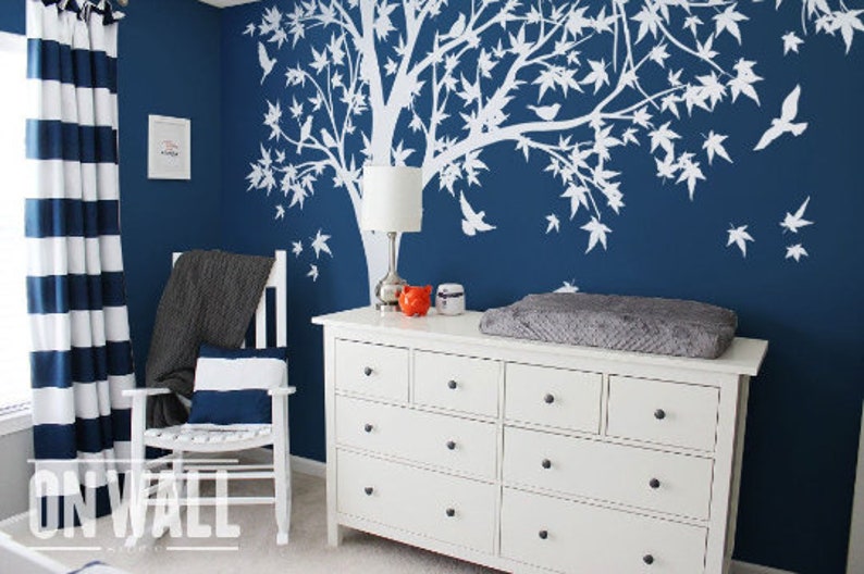 May include: White tree wall decal with birds on a dark blue wall, a white dresser with six drawers, a gray changing pad on top of the dresser, and a rocking chair with a gray throw blanket and a white and blue striped pillow.