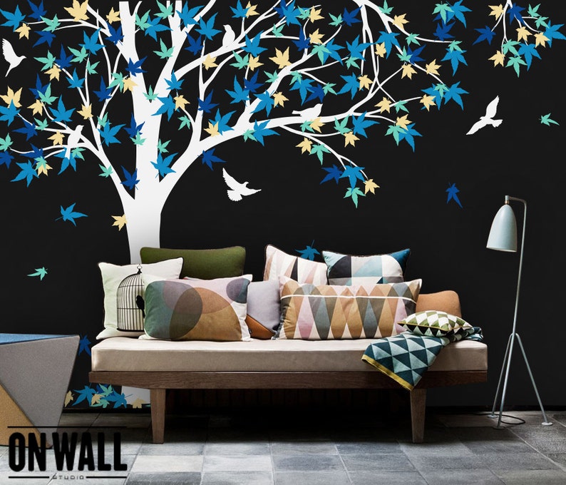 May include: A white tree silhouette with blue, green, and yellow leaves and small white birds on a black background. A couch with pillows and a throw blanket is in the foreground. The text "ON WALL Studio" is in the bottom left corner.