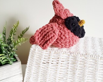 May include: A plush crochet bird toy featuring a rounded body made of soft pink yarn, a black face mask around the eyes, and a small yellow beak. The bird has a textured, fuzzy appearance with a prominent crest on top of its head.