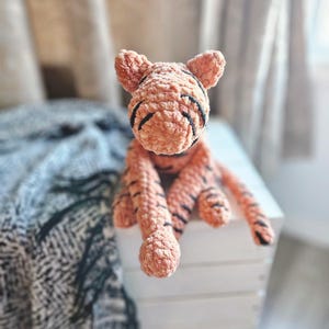 May include: A plush tiger toy crafted from soft, peach-colored chenille yarn. The stuffed animal features a rounded head with pointed ears, embroidered black stripes across its face and limbs, and long, dangling arms and legs with a textured finish.