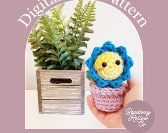 May include: A digital PDF pattern titled Petals the Little Flower featuring a crochet flower with a yellow face and blue petals in a light purple pot, next to a green fern in a wooden crate, with the Bearberry Hollow Co logo.
