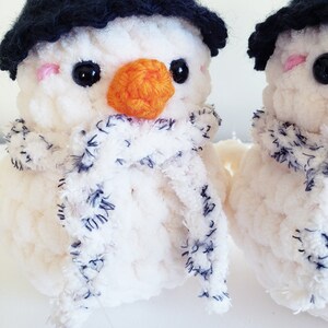 May include: Two white crocheted snowman ornaments with black hats and blue and white striped scarves. The snowman on the left has an orange felt nose.