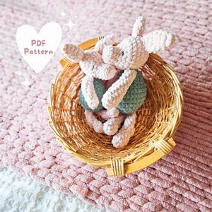 Könnte beinhalten: Zwei gehäkelte Schweine-Plüschtiere in einem Weidenkorb. Die Schweine-Plüschtiere sind rosa und eines hält ein grünes gehäkeltes Objekt. Der Korb befindet sich auf einer rosa gestrickten Oberfläche.
