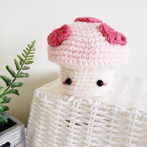 May include: A pink crocheted mushroom with red spots. The mushroom has a white stem and black eyes. It is sitting on a white wicker basket.