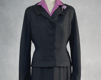 May include: A vintage black skirt suit with a matching jacket and knee-length skirt. The jacket features a purple collared shirt underneath and a decorative purple brooch. The suit is displayed on a mannequin.