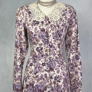 May include: A long-sleeved dress featuring a floral pattern in shades of purple and brown on a cream background. The dress has a button-down front and a large, cream-colored lace collar. A multi-strand pearl necklace is worn around the neck.