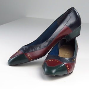 May include: A pair of multi-colored leather pumps. The shoes feature a color block design with navy blue, burgundy, and green panels. Gold-colored studs adorn the toe of each shoe. The heels are a low wedge style.