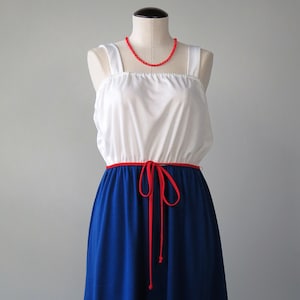 May include: A white sleeveless dress with a red ribbon tie at the waist and a blue skirt. The dress has a red beaded necklace.