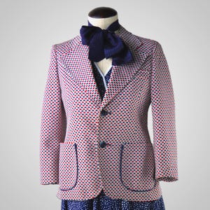 May include: A white blazer with a red and blue geometric pattern. The blazer has a navy blue bow tie and two pockets with blue trim.