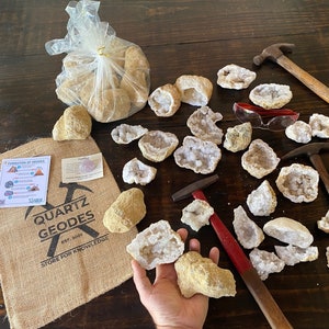 May include: A burlap bag with the text "Quartz Geodes Est. 2001 Store for Knowledge" and a diagram of the formation of geodes. A clear plastic bag filled with unopened geodes. A hand holding a broken geode revealing white crystals. Several broken geodes with white crystals on a wooden surface. Two hammers and safety glasses.