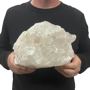 May include: A large, raw cluster of clear quartz crystals featuring multiple jagged, translucent points and facets. The mineral specimen displays a natural, irregular formation with varying degrees of transparency and white, cloudy inclusions throughout.