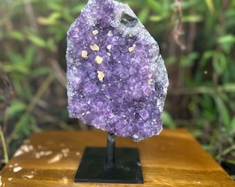 May include: A large, raw amethyst crystal cluster, showcasing deep purple hues and small, golden-yellow inclusions. The crystal is mounted on a black metal stand, set against a blurred green background, suggesting a natural setting. This decorative piece is ideal for home decor.