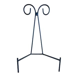 May include: A black metal display stand with a three-legged base and a decorative top featuring two curved swirls. The stand is designed to hold plates or artwork and is suitable for home decor.