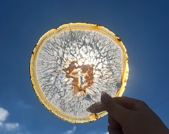 Könnte beinhalten: Eine transparente, kreisförmige Agatscheibe mit hellbraunem und weißem Muster, gehalten vor einem strahlend blauen Himmel. Der Agat hat einen goldbraunen Außenrand und komplizierte innere Aderung.