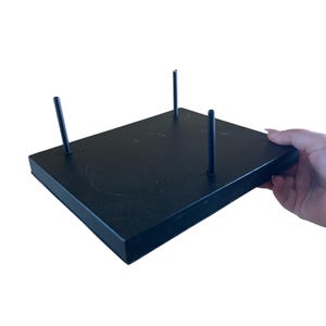 May include: A black rectangular display stand with three vertical rods. The stand is held by a hand, and the background is white. The stand is likely used for displaying items.