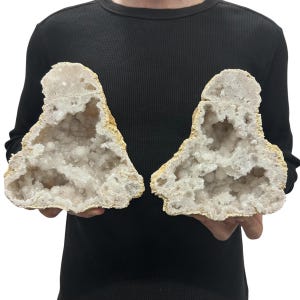 May include: Two large, matching halves of a geode featuring a rough, tan-colored exterior and a hollow interior filled with sparkling, translucent white quartz crystals. The mineral specimens have an irregular, jagged shape and texture.